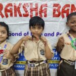 Three smiling schoolchildren in beige uniforms display handmade bead bracelets at a school event with a banner backdrop on the wall